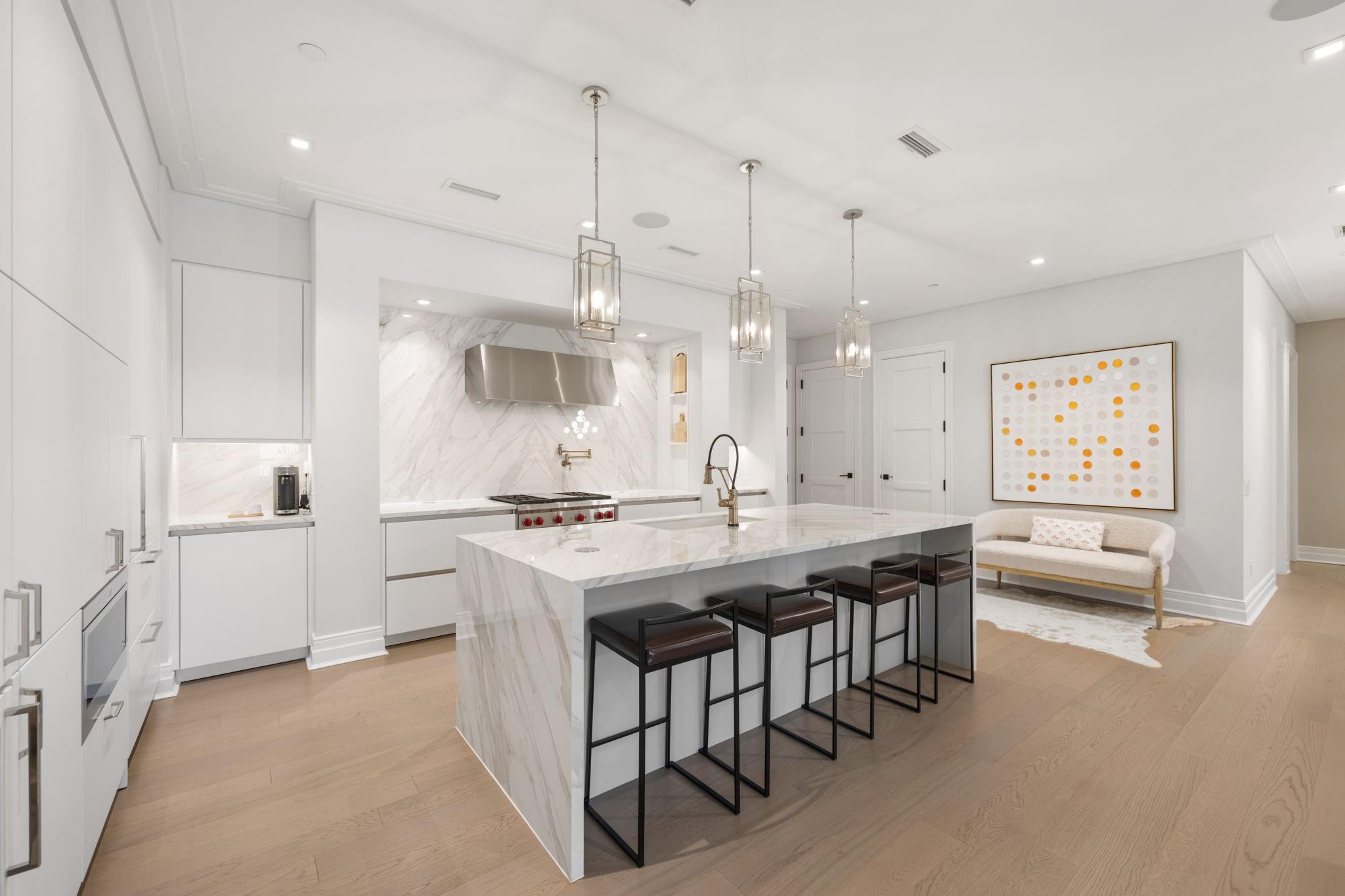 Modern white kitchen with a marble island, brass faucet, dark bar stools, and pendant lights.