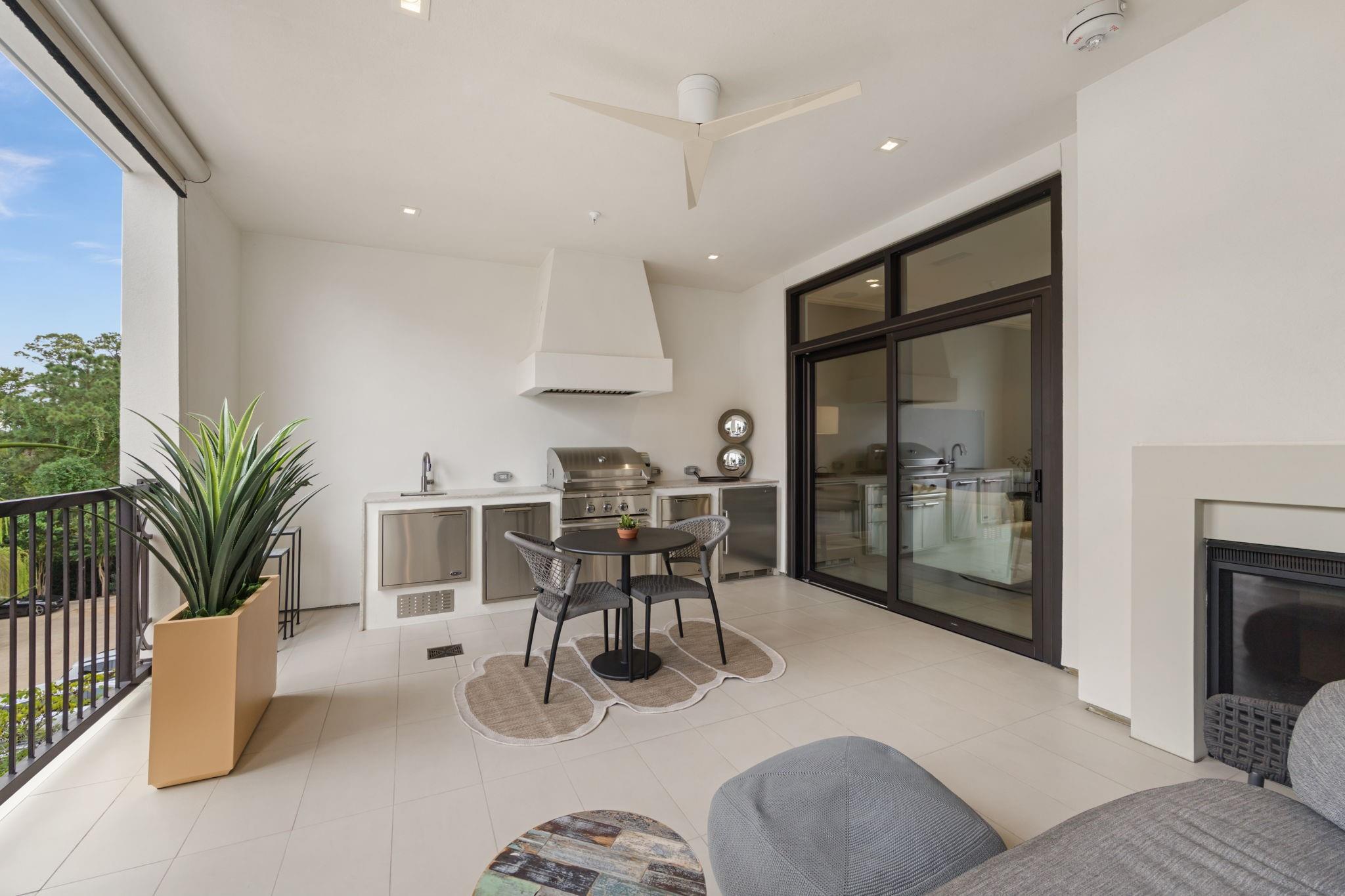 Covered terrace with a stainless outdoor kitchen, dining set, and fireplace beside sliding glass doors.