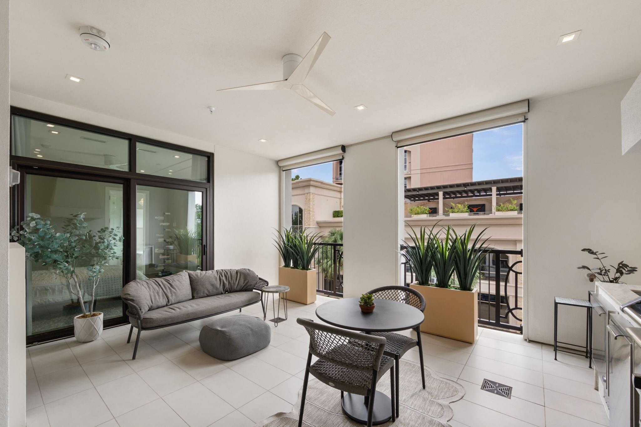 Furnished apartment balcony with a gray sofa, round table, and large potted plants.
