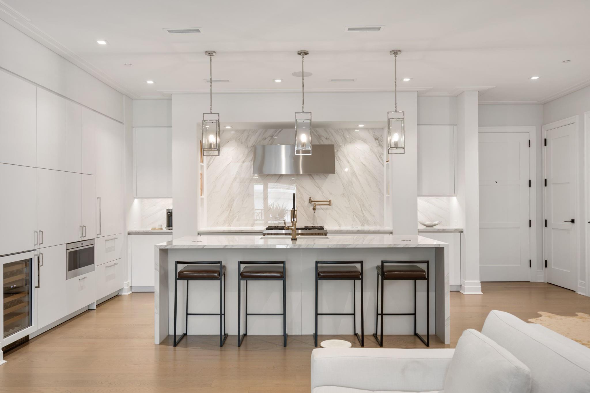 Modern white kitchen with a marble island, four bar stools, and pendant lights.