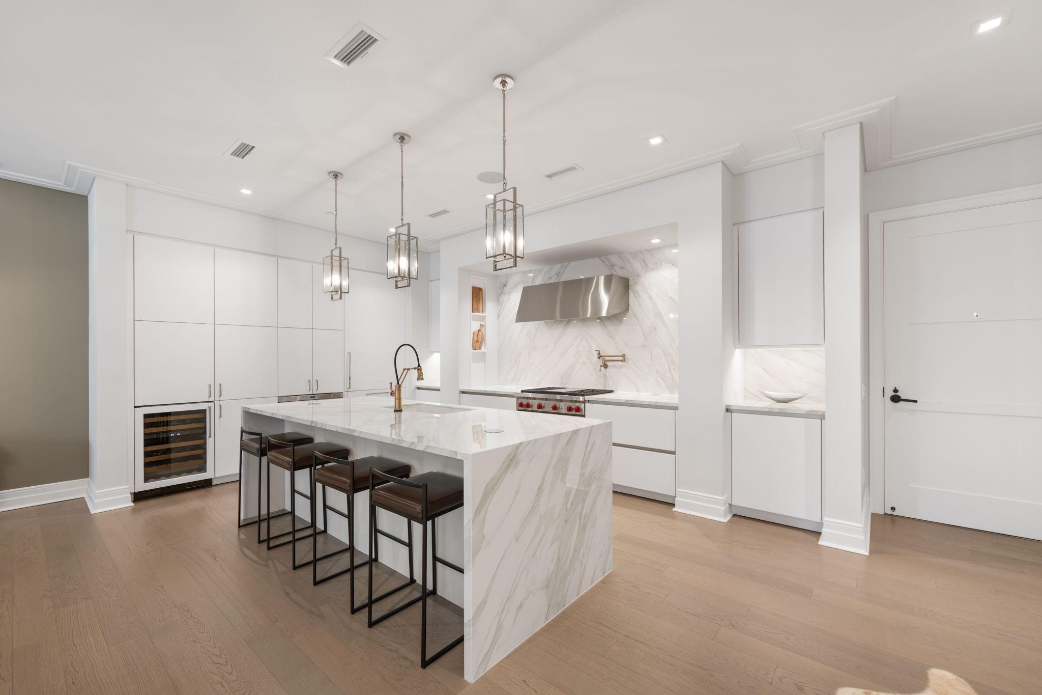 Modern white kitchen with a marble island, four bar stools, brass fixtures, and pendant lights.