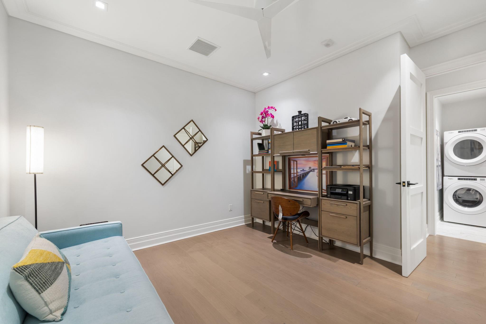 Home office with built-in wood desk and shelving, blue sofa, and adjoining laundry area.
