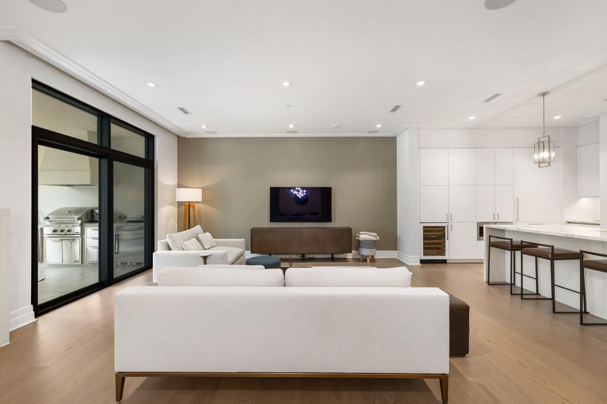 Open-plan living room with white sofas, a wall-mounted TV, kitchen island, and sliding doors to an outdoor grill.