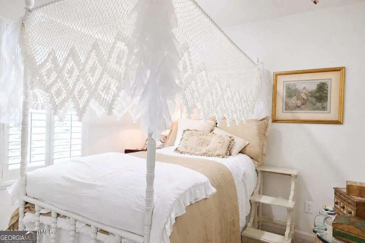 Bedroom with a white lace-draped four-poster bed and beige bedding beside a bright window.