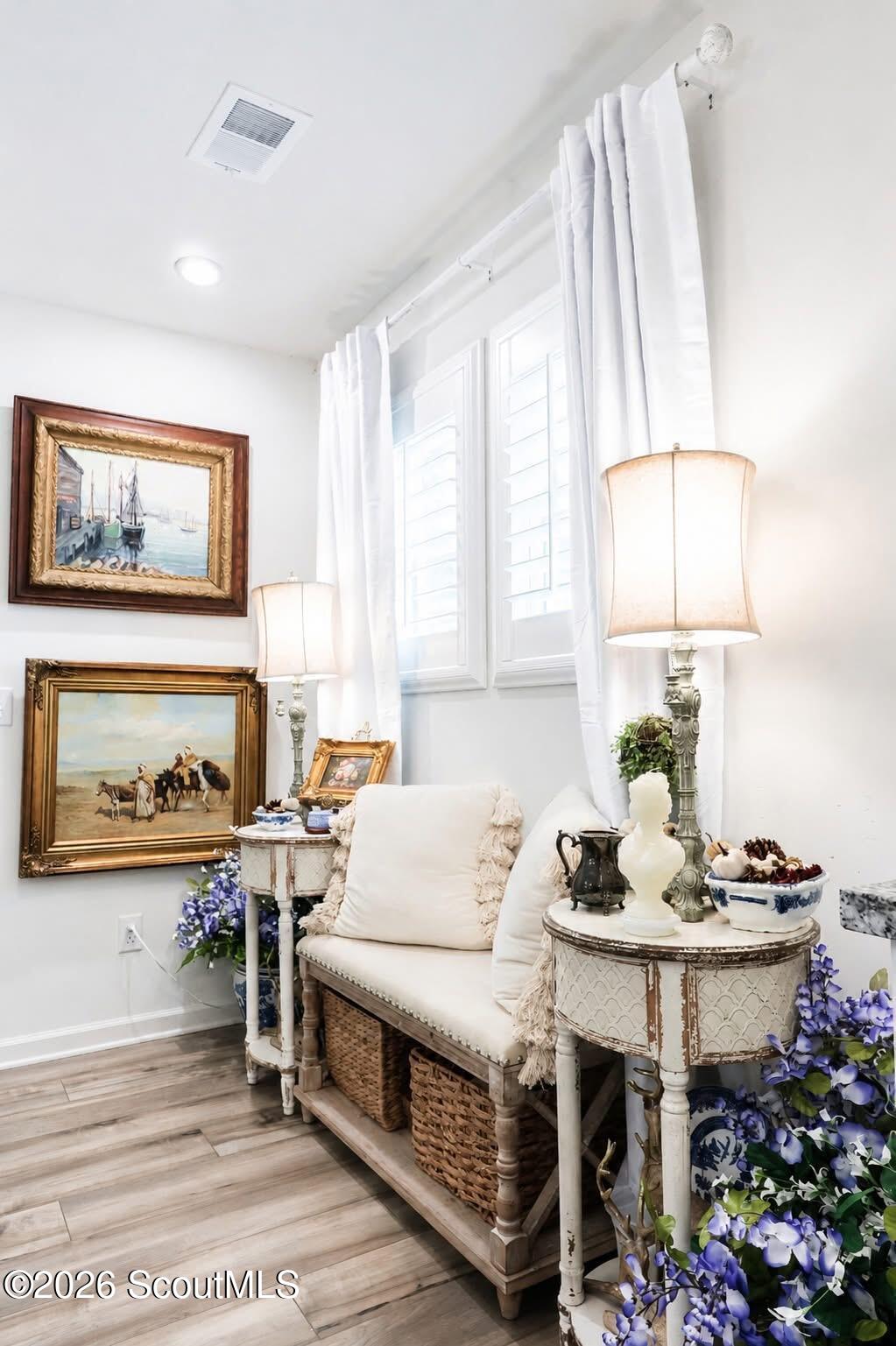 Sitting nook with a white cushioned bench, shuttered windows, ornate tables, framed art, and purple flowers.
