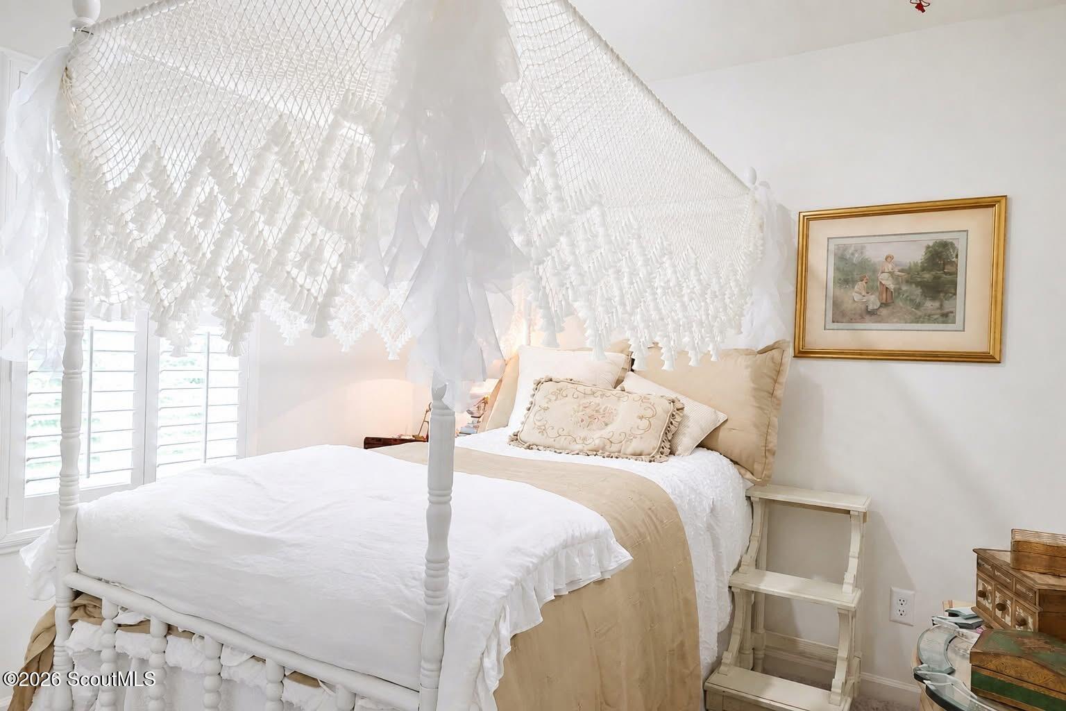 Bedroom with a white net-draped canopy bed, cream bedding, plantation shutters, and framed art.
