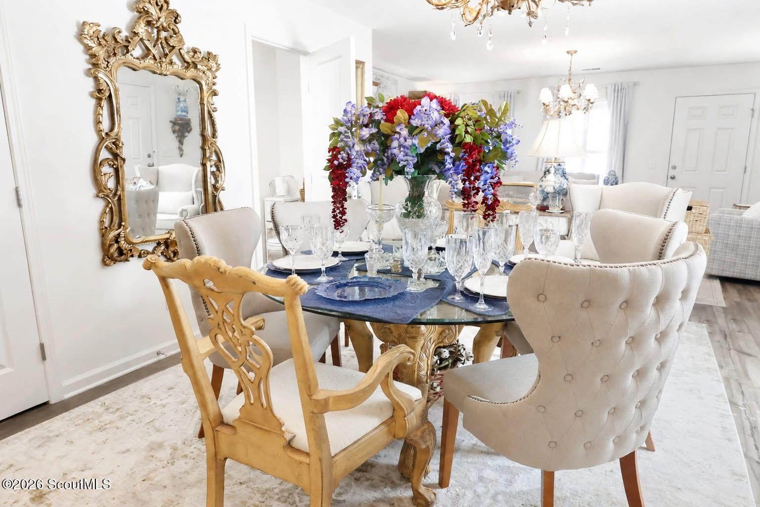 Dining room with a glass-top table, colorful floral centerpiece, crystal stemware, and ornate gilded mirror.