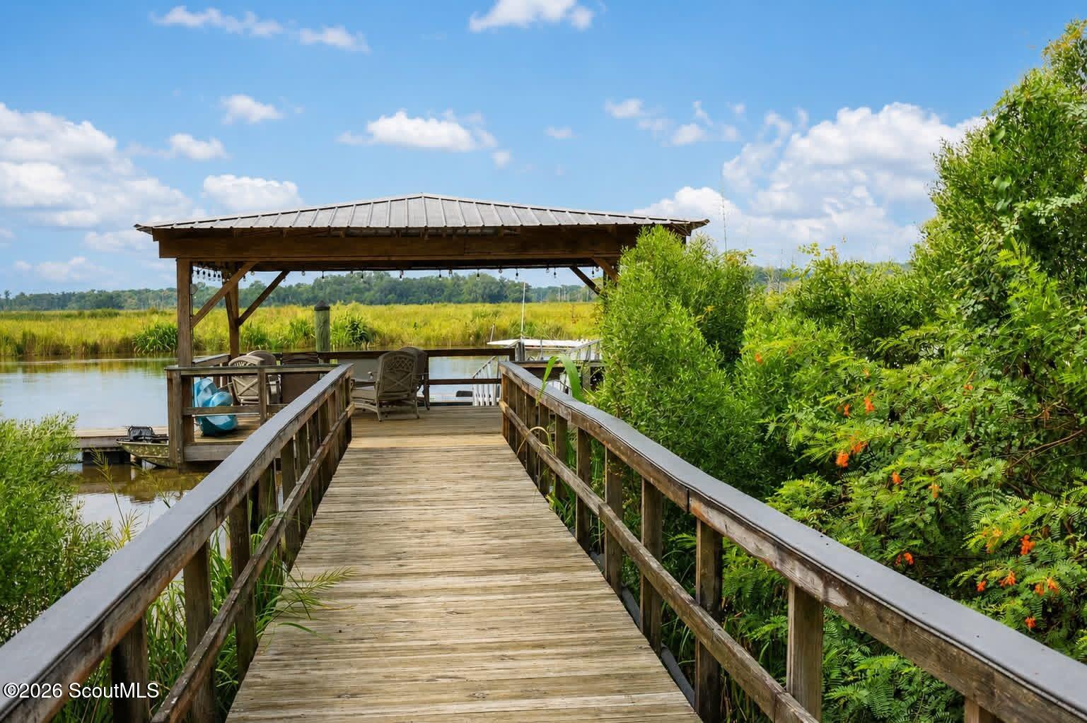 Wooden boardwalk leads to a covered waterfront dock, with marsh grasses and water beyond.
