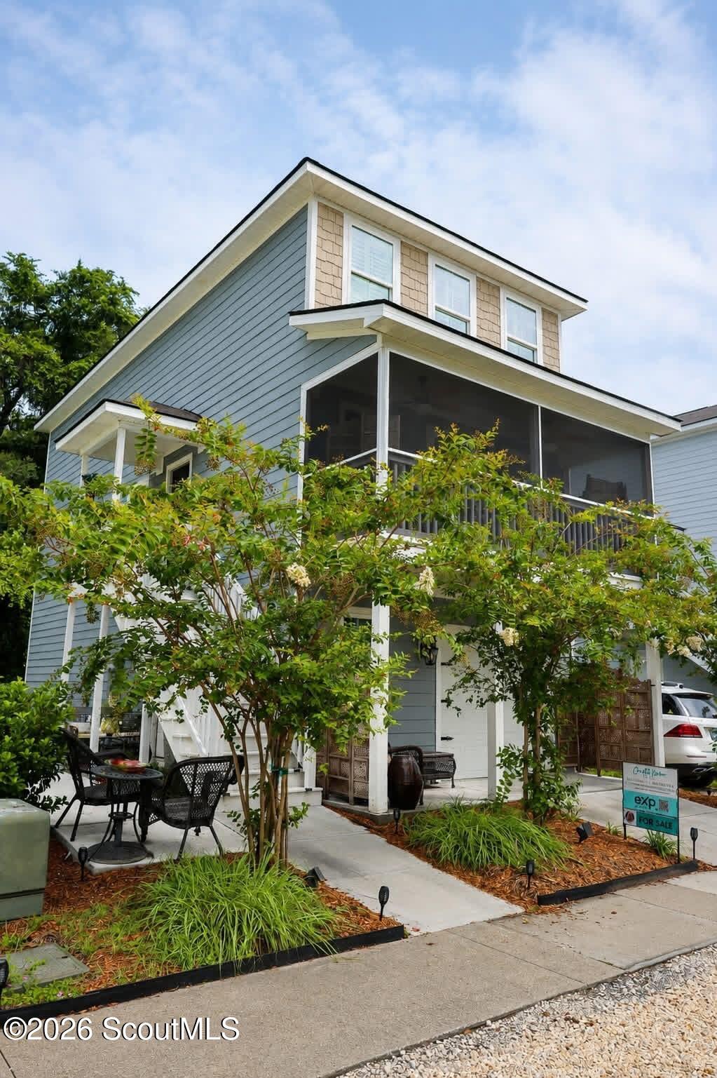 Blue-gray multi-level house with a screened porch, leafy landscaping, and a patio seating area.