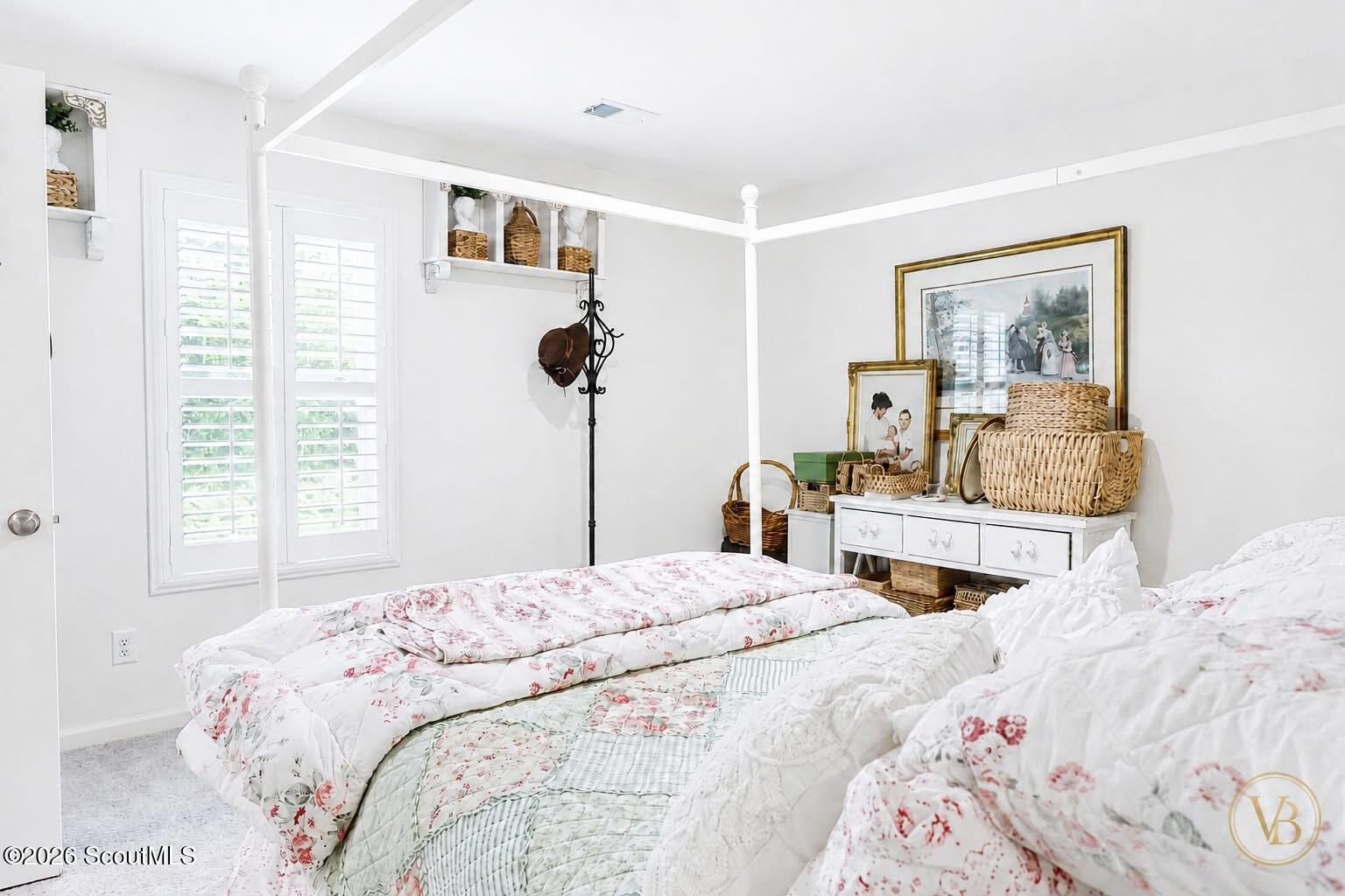 Bedroom with a white canopy bed, floral quilts, plantation shutters, and wicker baskets.