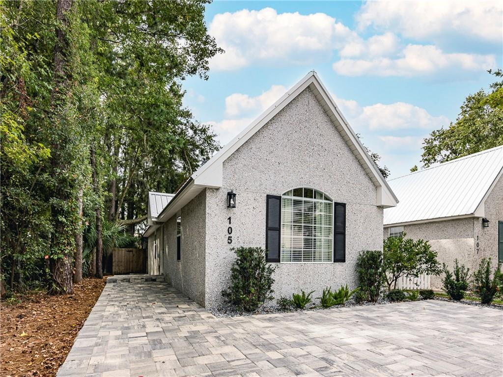 Gray stucco home with a steep gable, black shutters, and a paved driveway beneath trees.