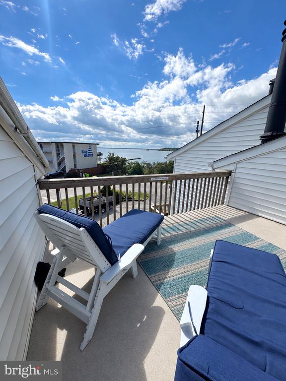 Rooftop deck with blue-cushioned lounge chairs, a striped rug, and a glimpse of water.