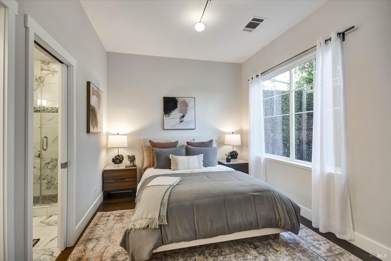 Staged bedroom with a gray-covered bed, large curtained window, and doorway to a tiled bathroom.