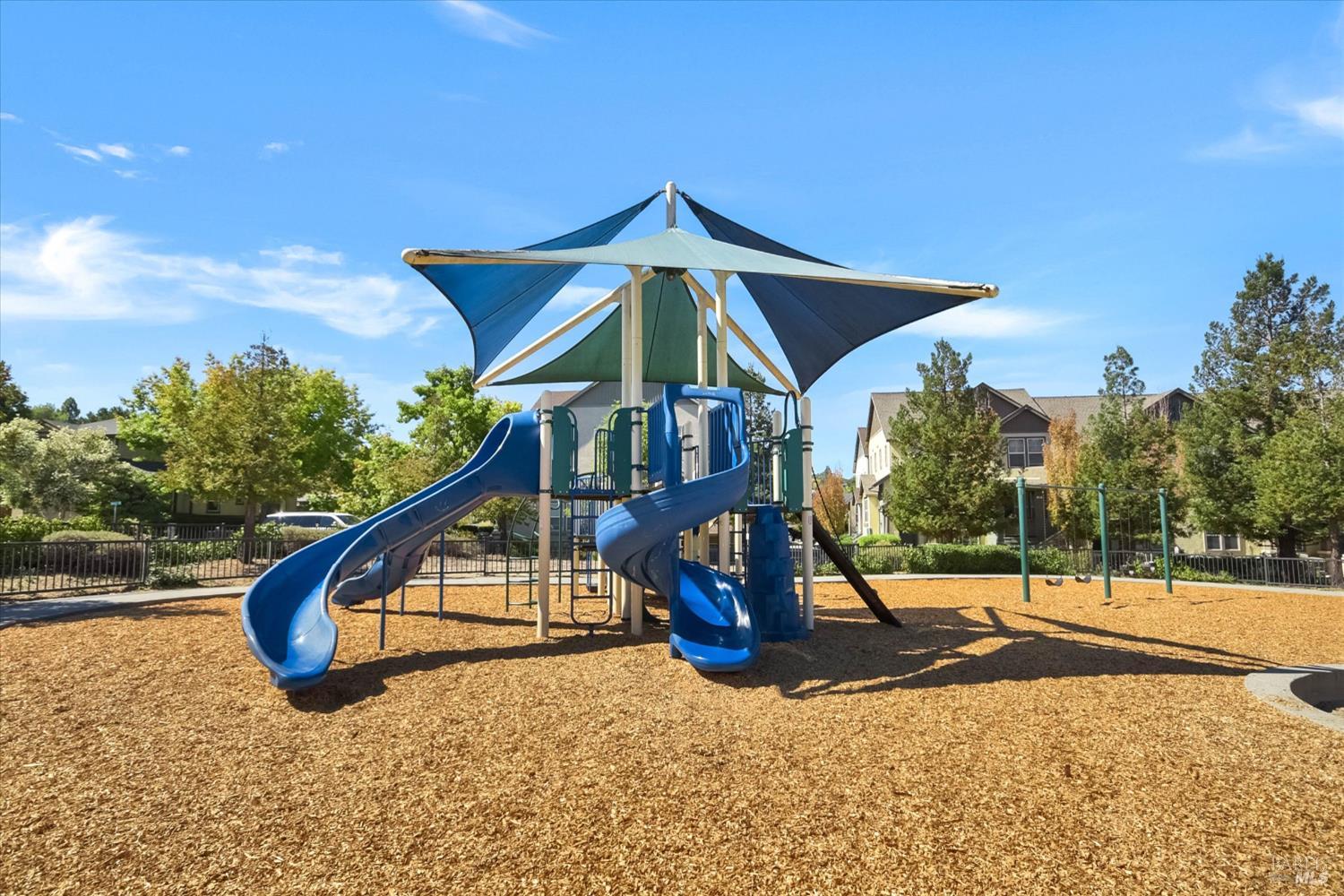 Community playground with blue slides and a large shade canopy over wood-chip surfacing.