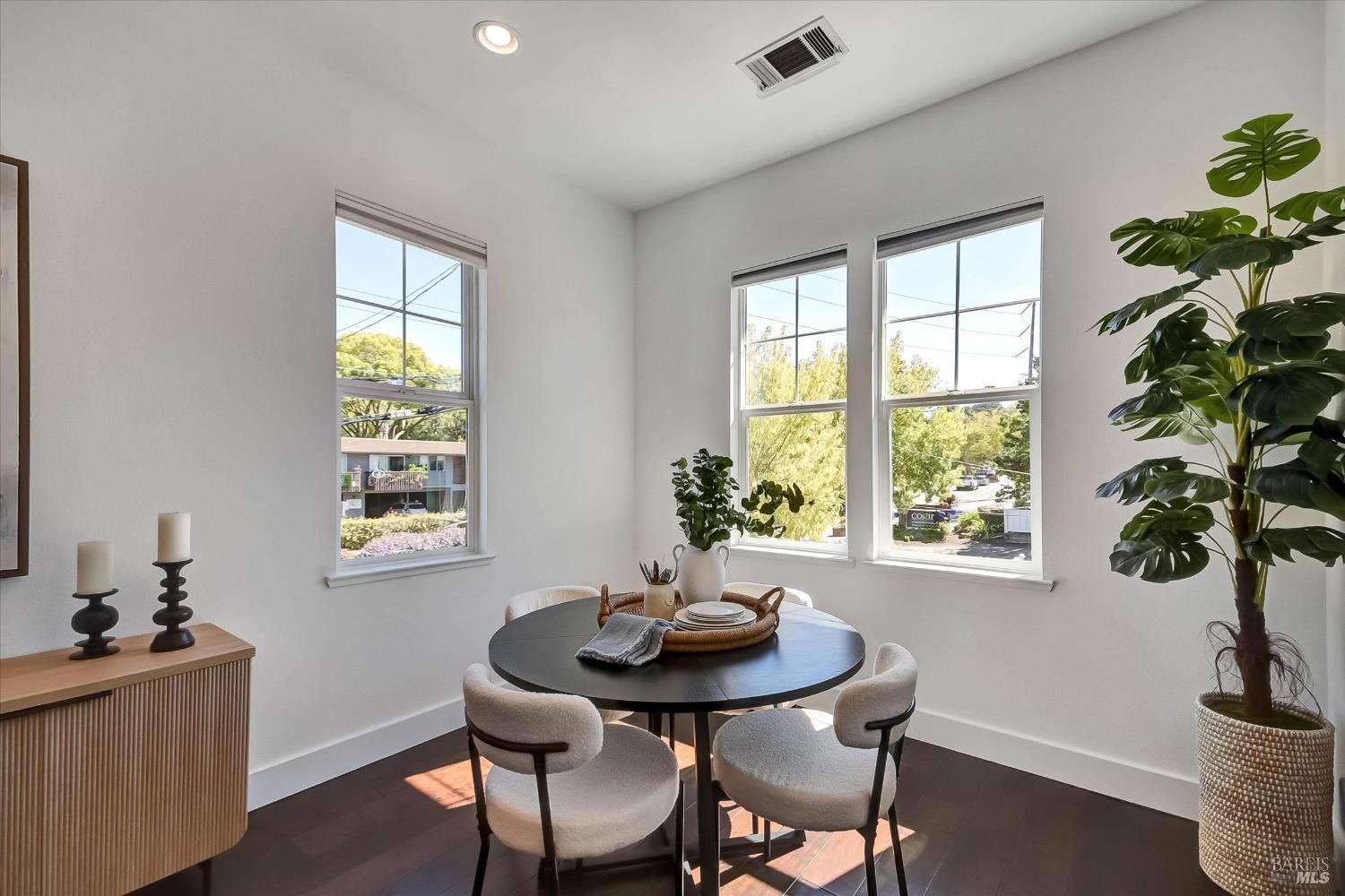 Dining area with a round black table, upholstered chairs, and three sunny windows.