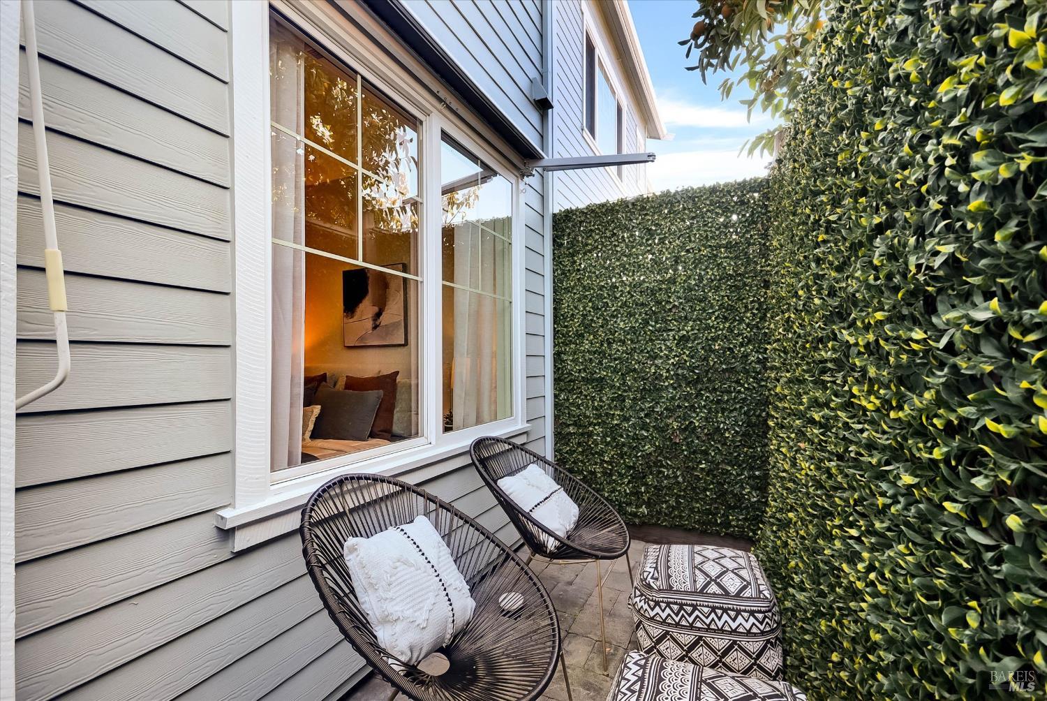 Private patio with woven chairs and patterned cushions, enclosed by tall hedges beside the home.
