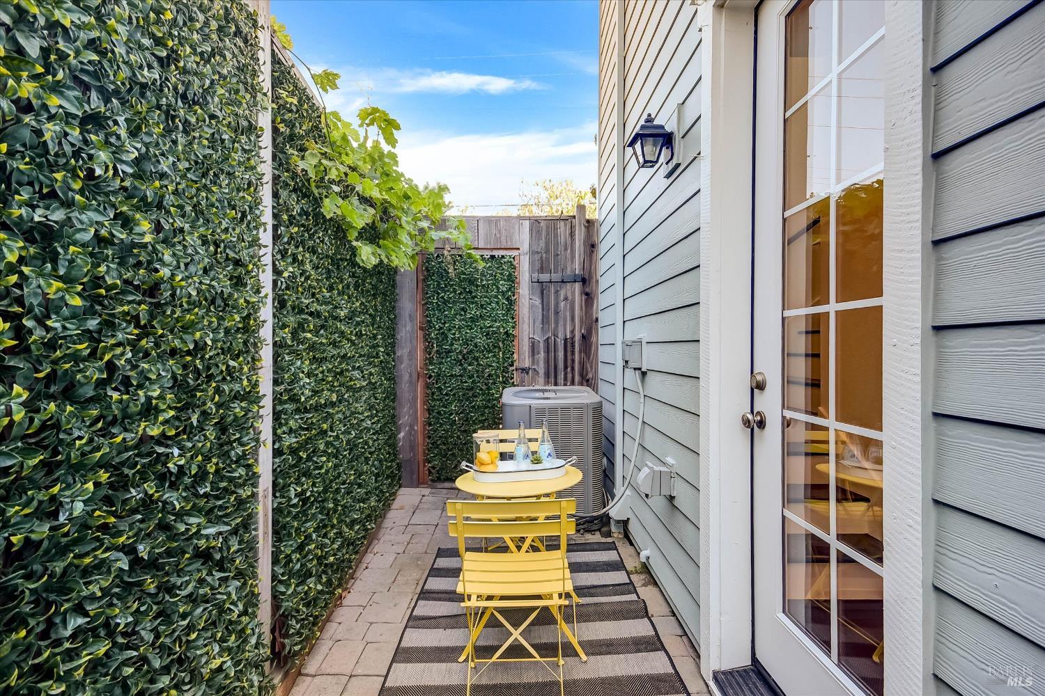 Narrow patio with tall hedges, a yellow bistro set, and a glass-paneled house door.