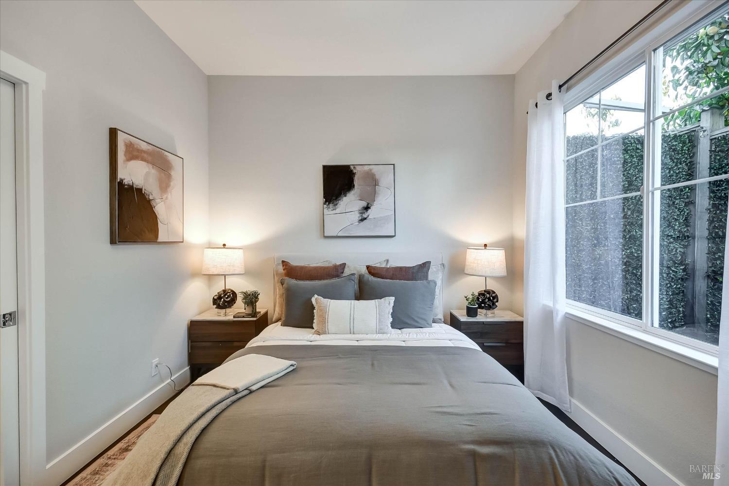 Staged bedroom with a gray-and-white bed, matching bedside lamps, and a large window beside a privacy hedge.