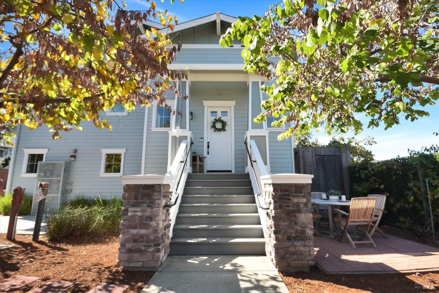 Blue-gray home entrance with a broad staircase, stone pillars, and a patio beneath autumn foliage.