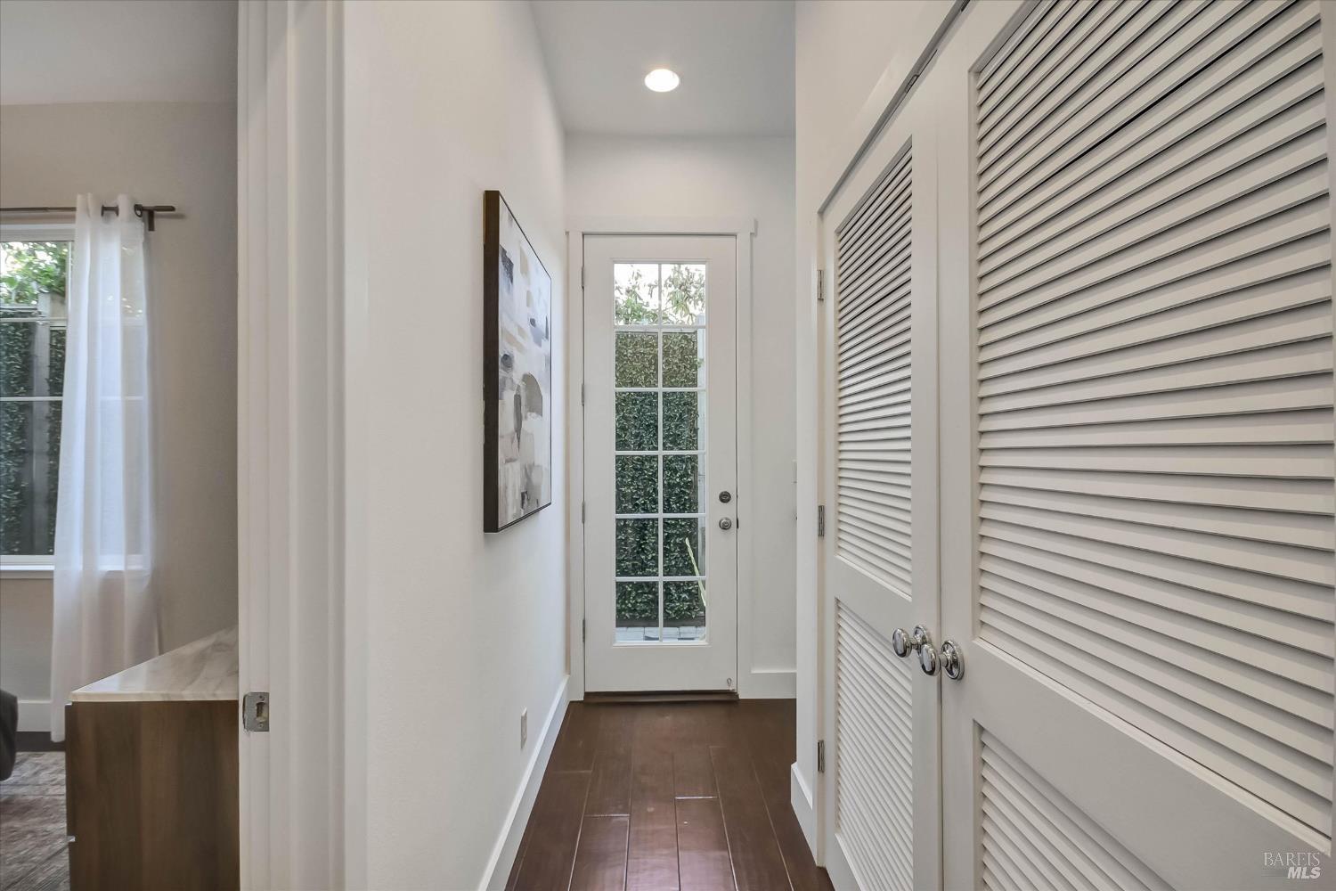 Hallway with dark wood floors, louvered closet doors, and a glass-paneled door opening toward a leafy hedge.