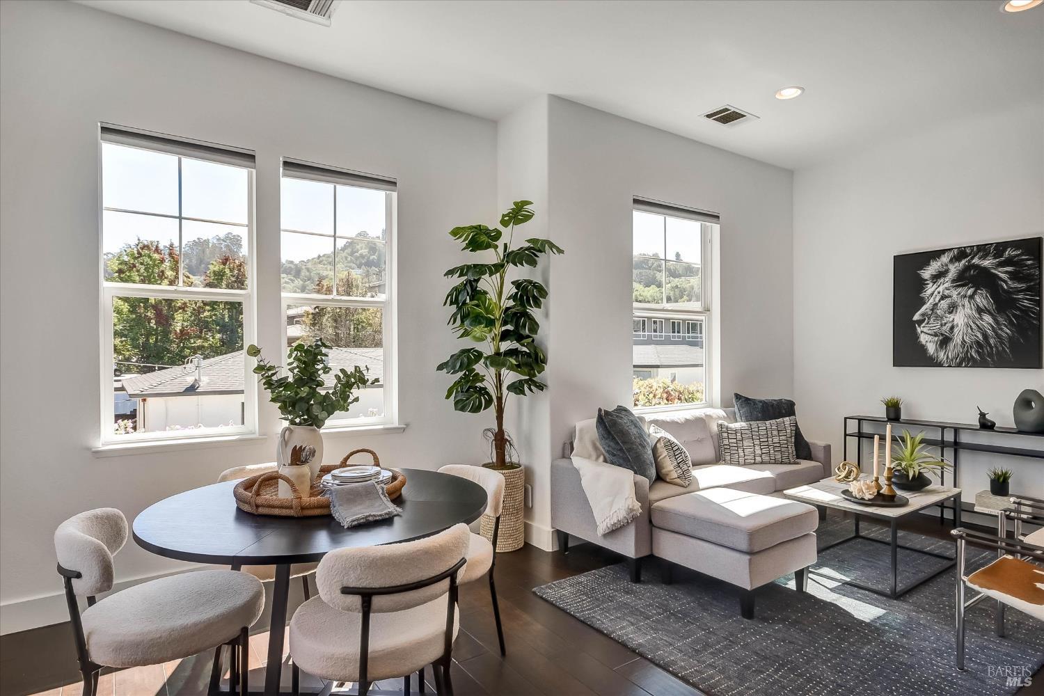Staged living and dining area with a gray sectional, round table, and large windows overlooking wooded hills.