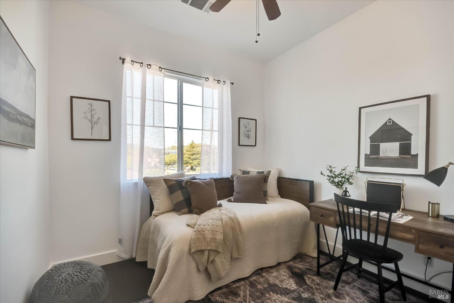 Staged bedroom with a curtained window, neutral bed, and desk with chair.