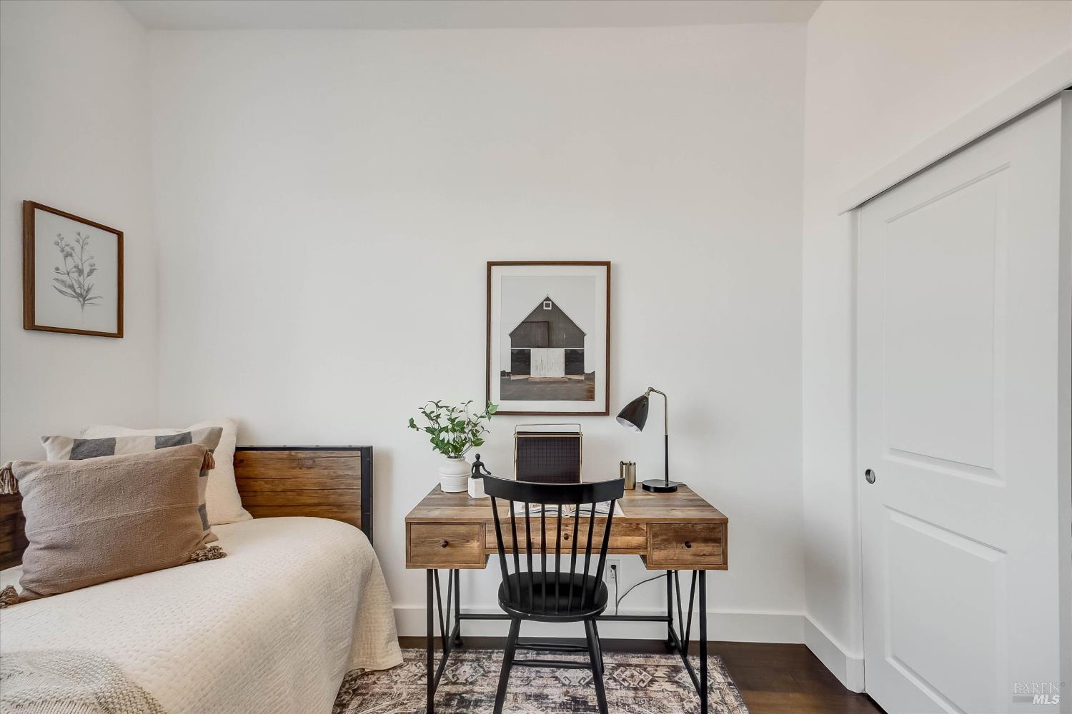 Staged bedroom with a white bed and wood desk beneath framed prints.