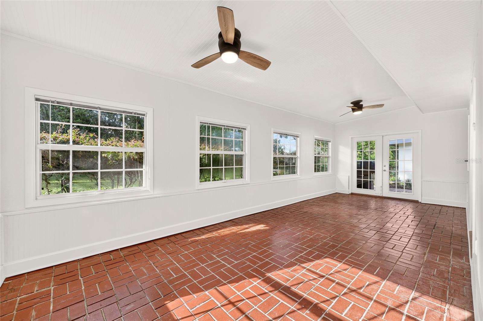 Enclosed sunroom with red brick floors, bright windows, and two ceiling fans.