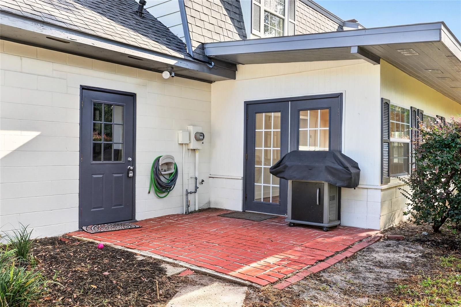 Rear patio with brick paving, French doors, and a covered grill beside a white-sided house.