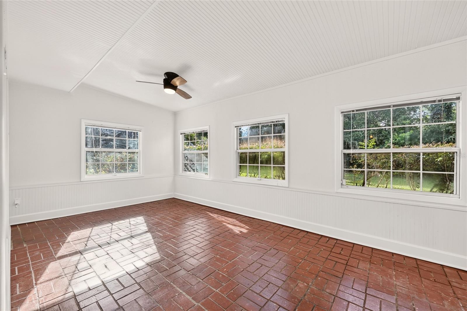 Empty sunroom with a red brick floor, white beadboard walls and ceiling, and four windows overlooking greenery.