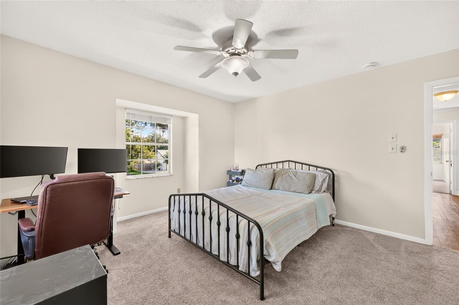 Bedroom with a black metal bed, ceiling fan, and desk with two monitors beside a window.
