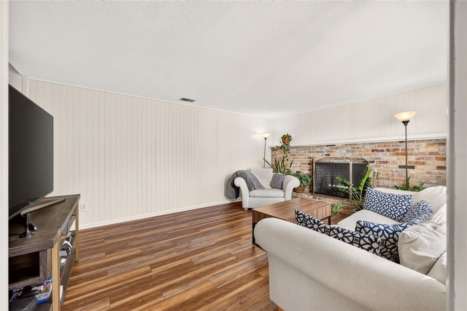 Living room with a brick fireplace, white seating, wood-look floors, and a television.
