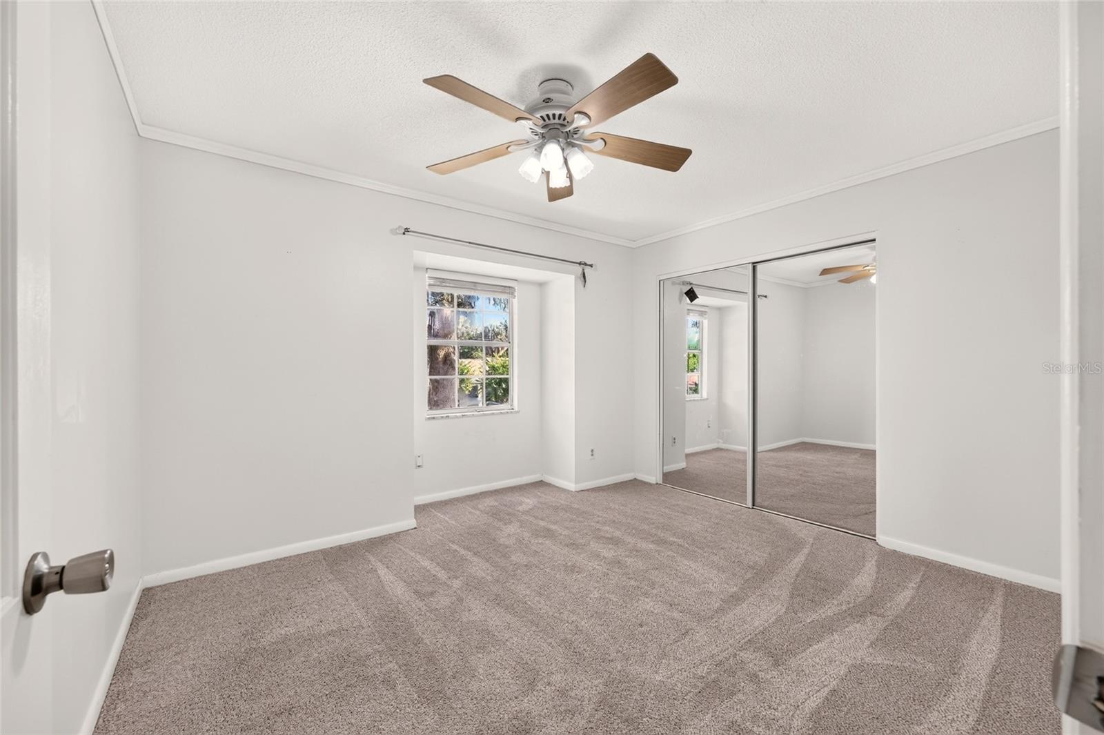 Vacant bedroom with gray carpet, mirrored closet doors, a window, and a ceiling fan.