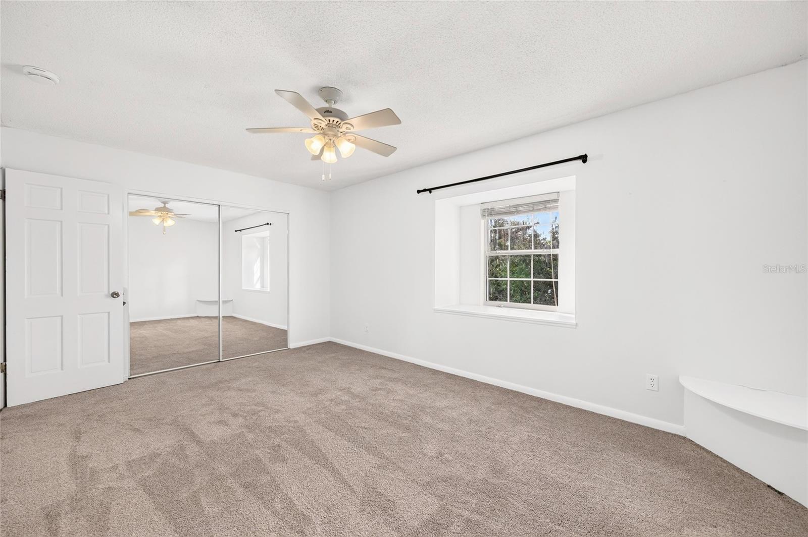 Empty bedroom with beige carpet, mirrored closet doors, and a ceiling fan.
