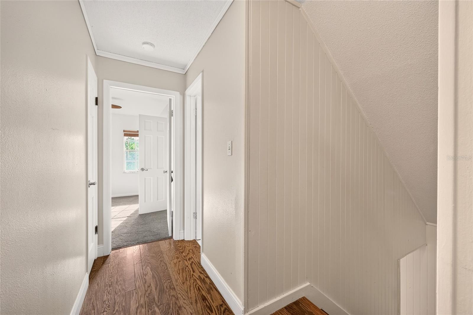 Hallway with wood floors beside a paneled stairwell and an open doorway to a carpeted room.
