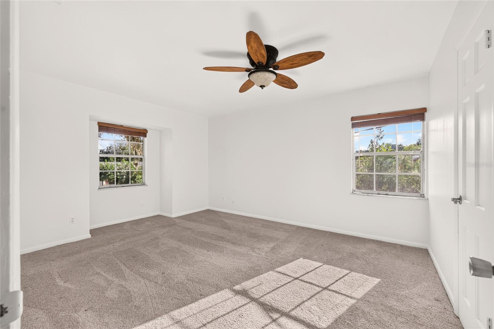 Empty bedroom with two windows, gray carpet, and a wood-blade ceiling fan.