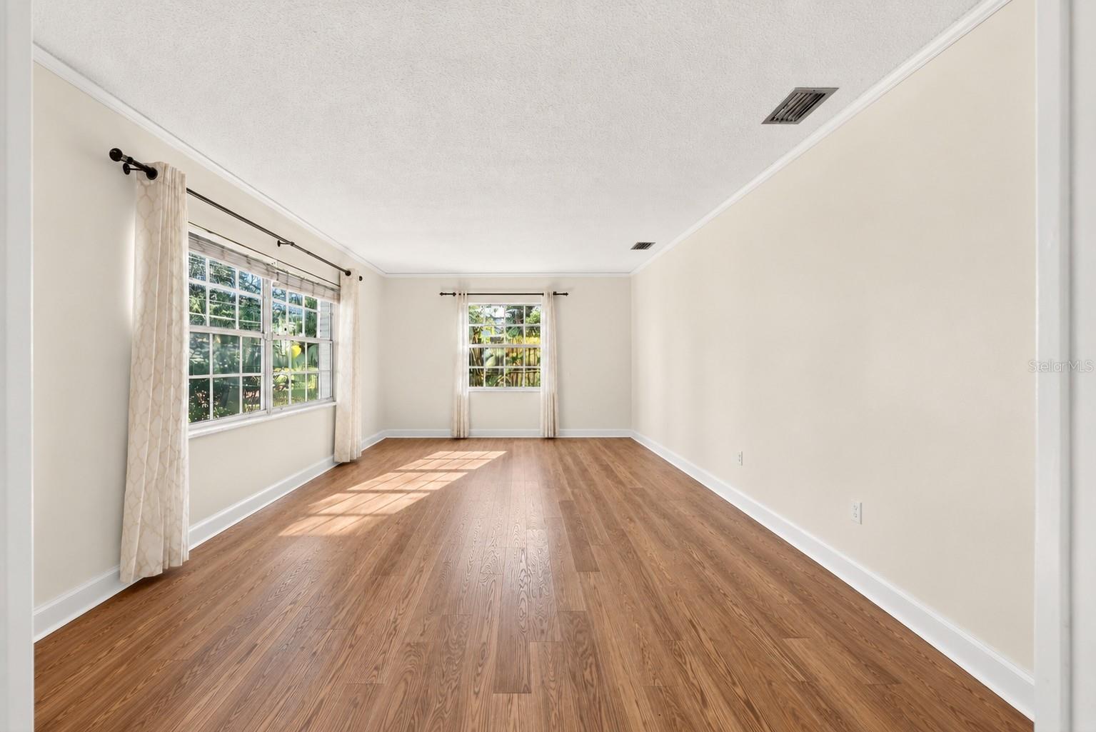 Empty room with warm wood flooring and large curtained windows.