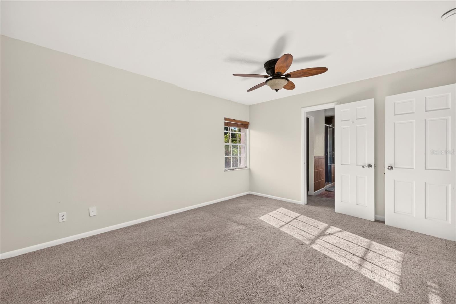 Empty bedroom with gray carpet, a ceiling fan, and sunlight streaming through the window.