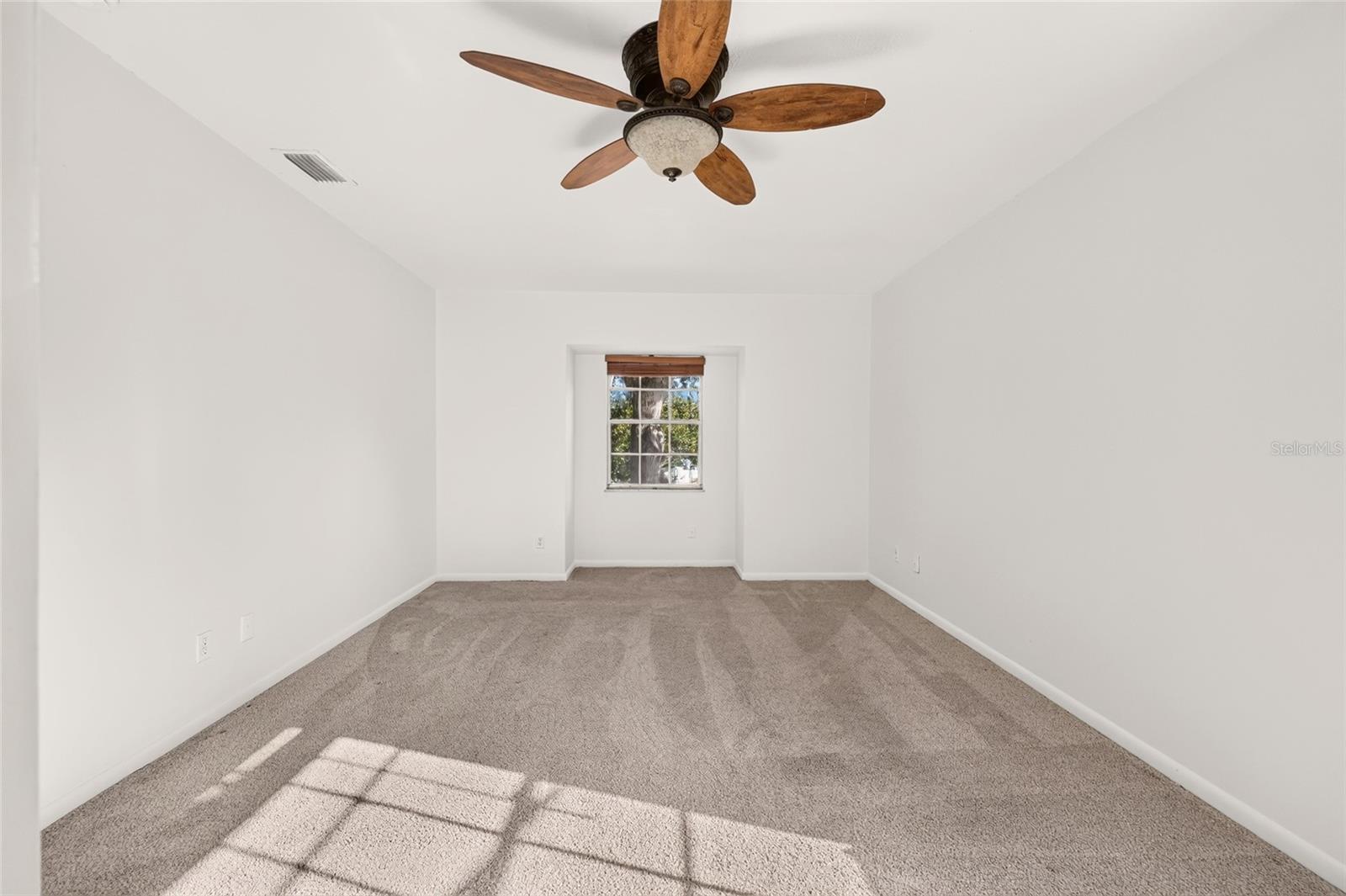 Empty bedroom with beige carpet, a wood-blade ceiling fan, and sunlight streaming through the window.