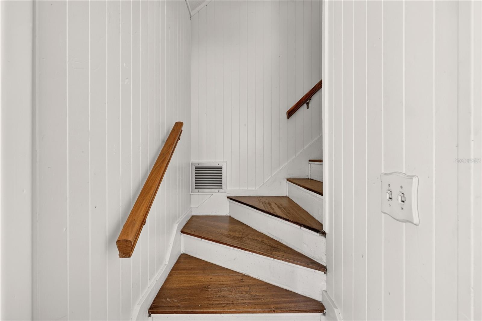 Interior staircase with worn wooden treads, wooden handrails, and white paneled walls.