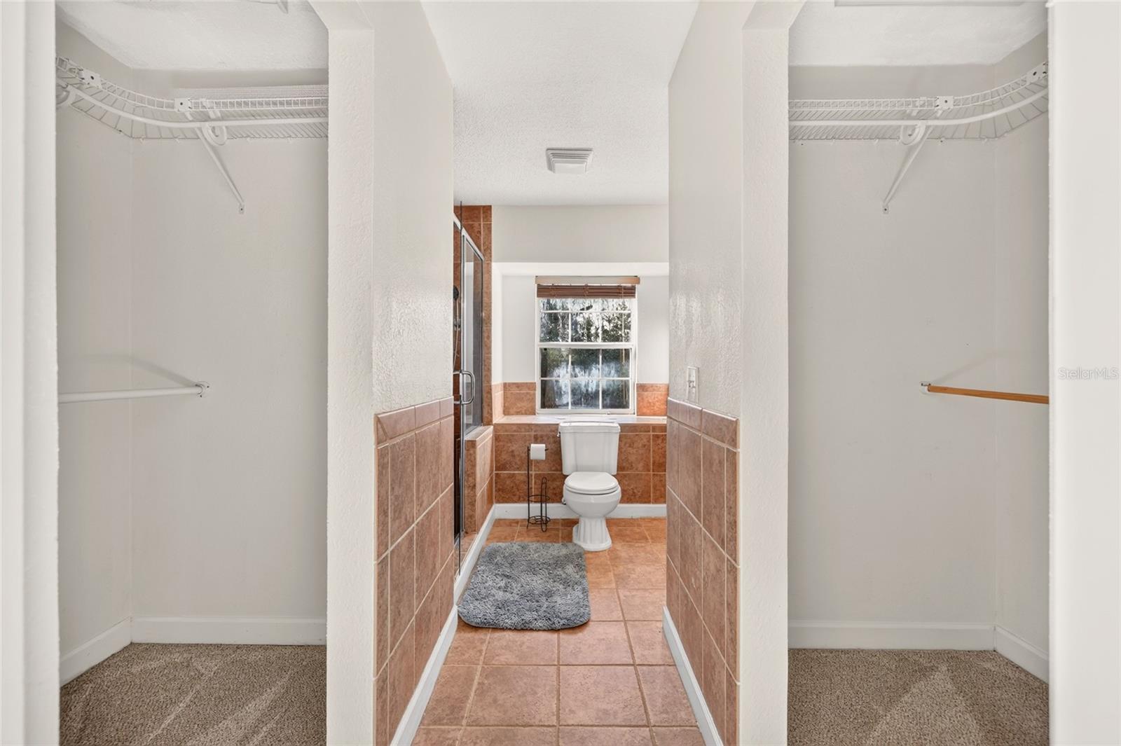 Bathroom with terracotta tile, a toilet beneath a window, and open closets on both sides.