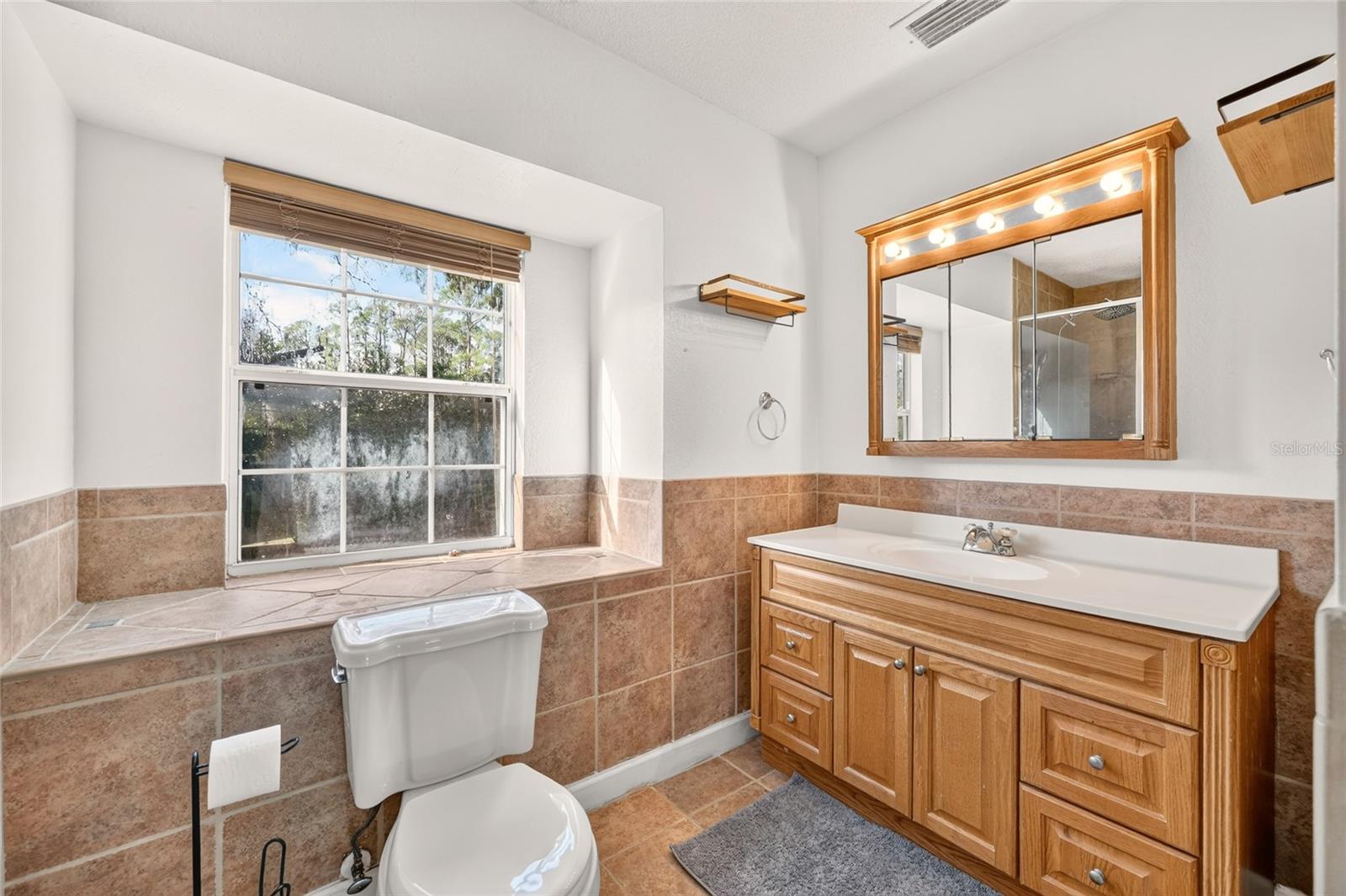 Bathroom with a wood vanity, illuminated mirror, tan tile and a large window.