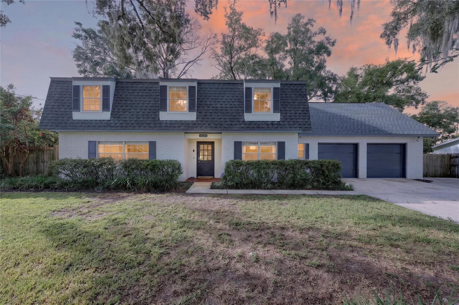 White-brick house with three dormers, dark shutters, and a two-car garage at dusk.