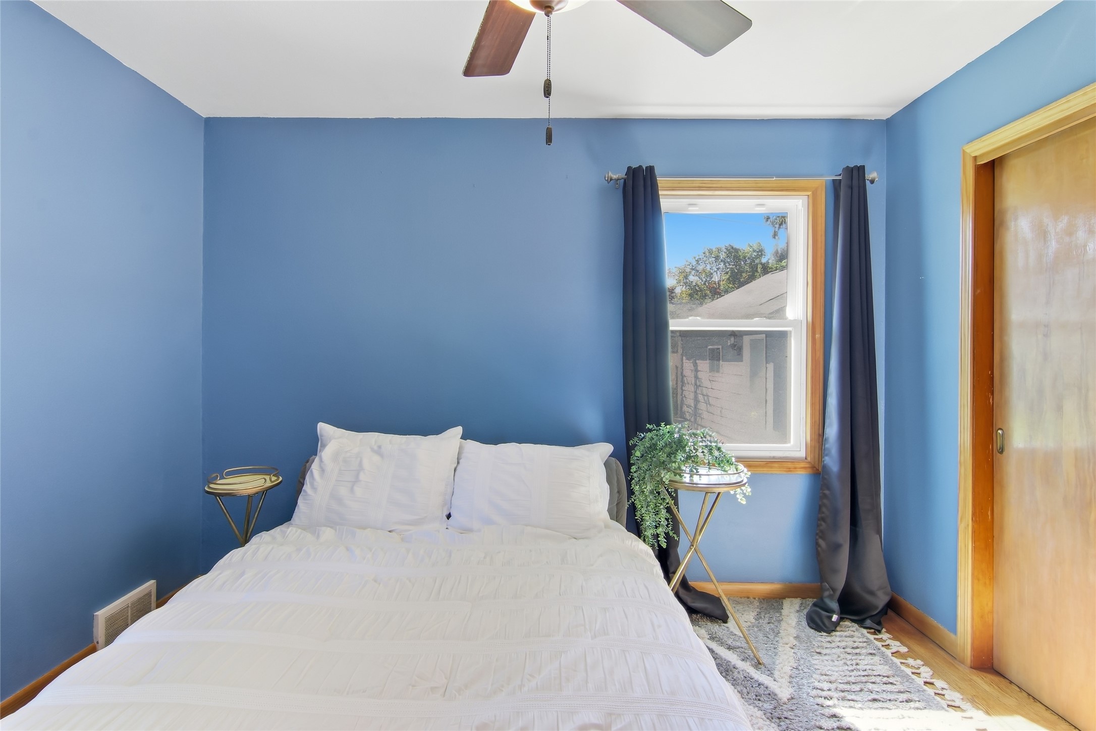 Bedroom with blue walls, a white bed, dark curtains, and a wood-trimmed window.