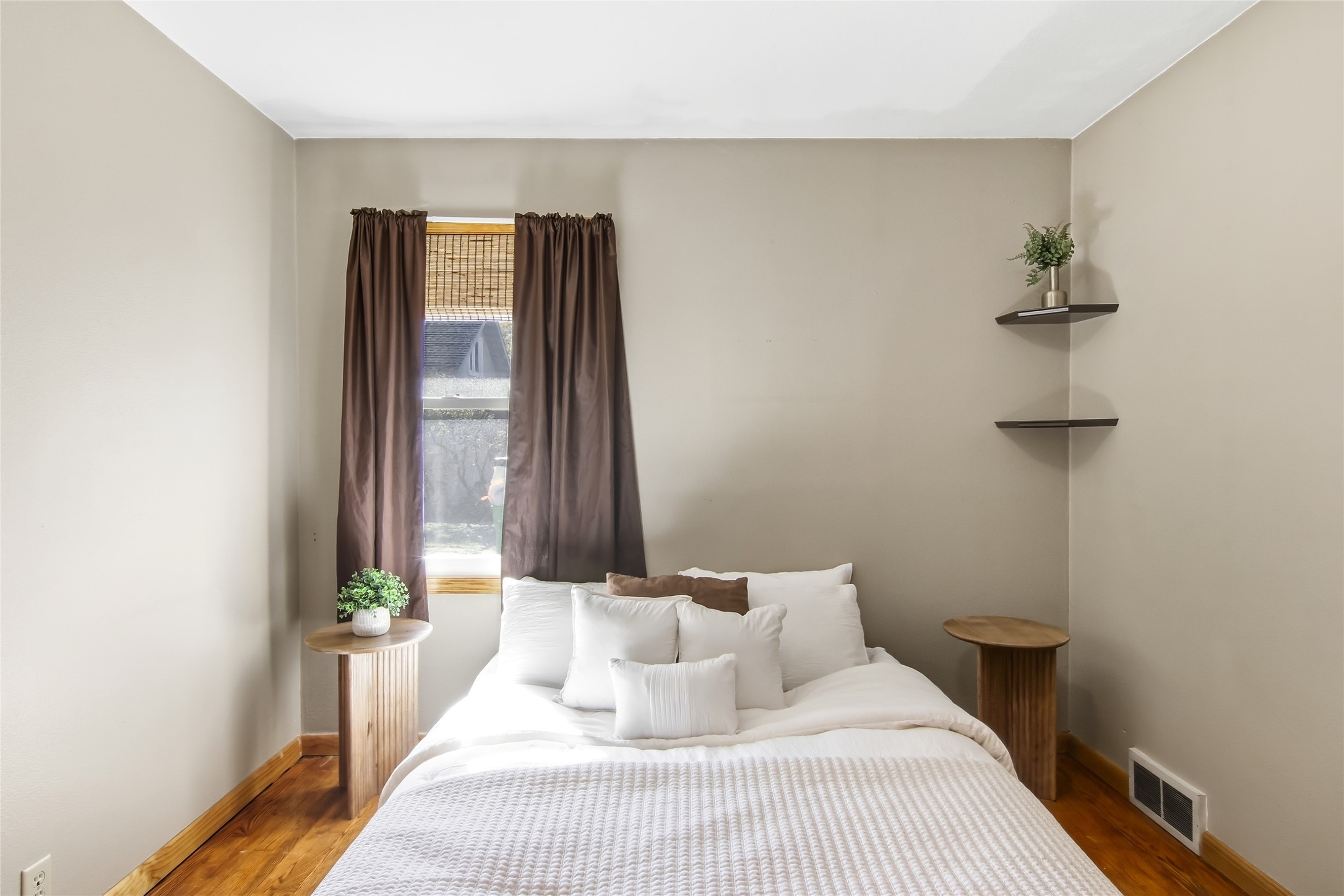 Staged bedroom with a white bed, dark window curtains, and warm wood flooring.