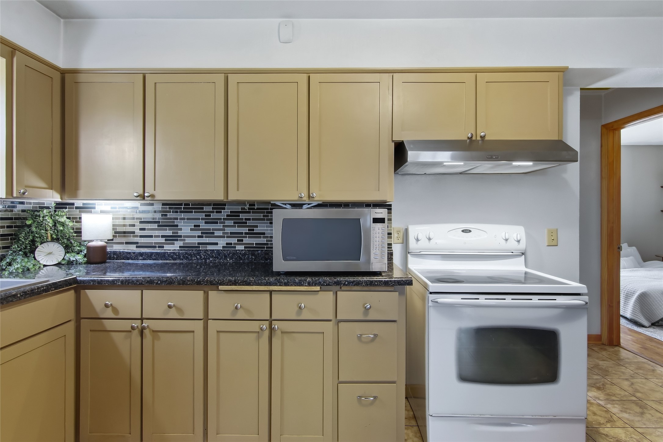 Kitchen with tan cabinets, dark counters, black tile backsplash, and white range beneath a stainless-steel hood.