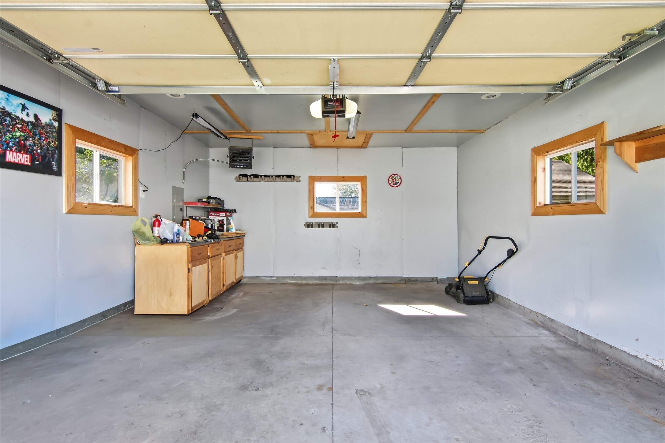 Garage with open overhead door, wood cabinets, three windows, and a lawn mower.