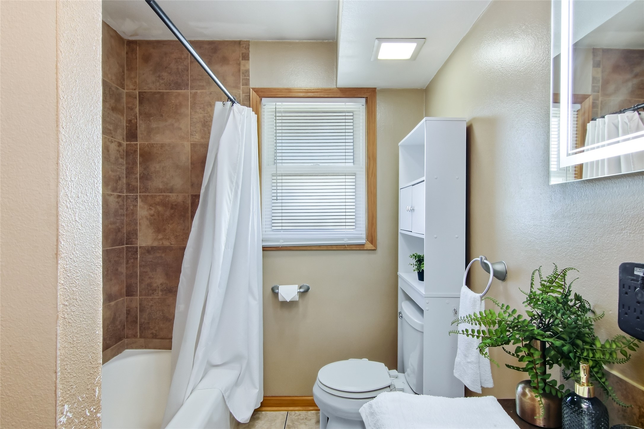 Bathroom with brown tiled tub surround, white shower curtain, toilet, and window with blinds.