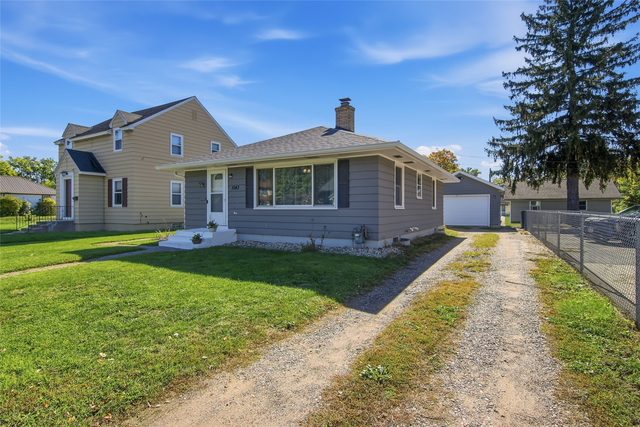 Gray single-story home with a wide picture window, white entry steps, and gravel driveway to a detached garage.