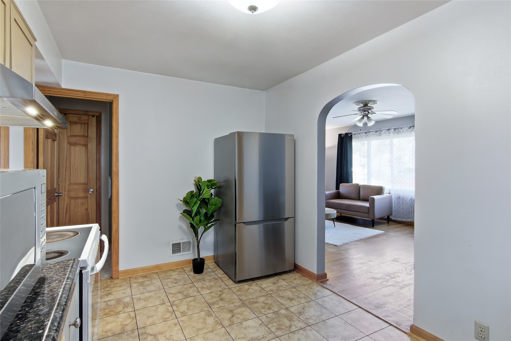 Kitchen with stainless steel refrigerator and tiled floor, opening through an arch to a bright living room.